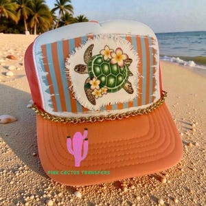 May include: A coral trucker hat with a white top and mesh back. The front has a patch with a green turtle and flowers, set against a striped background. A gold chain trims the brim, and a pink cactus is on the bill. The text "PINK CACTUS TRANSFERS" is at the bottom.