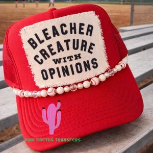 May include: A red trucker hat with a white patch that reads "BLEACHER CREATURE WITH OPINIONS." The hat features a pink cactus graphic and a beaded embellishment. The text "PINK CACTUS TRANSFERS" is printed on the brim.
