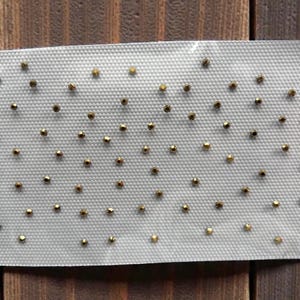 Könnte beinhalten: Eine weiße, strukturierte Folie mit kleinen, goldfarbenen Nieten. Die Nieten sind gleichmäßig verteilt und scheinen dekorative Elemente zu sein, möglicherweise für Bastelarbeiten oder Modeaccessoires. Der Hintergrund ist Holz.