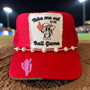 Puede incluir: Gorra de béisbol roja con parte trasera de malla y visera curva. La parte delantera presenta un parche con el texto "Take me out to the Ball Game" y un personaje de béisbol. Un cactus rosa y cuentas temáticas de béisbol adornan la gorra.