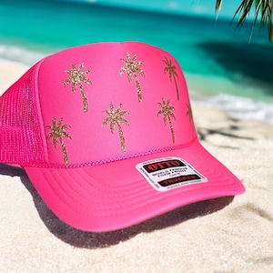 Könnte beinhalten: Hellrosa Trucker-Cap mit Mesh-Rückseite und gebogenem Schirm. Auf der Vorderseite des Hutes befinden sich goldene Glitzer-Palmenmuster. Der Hut liegt auf einem Sandstrand.