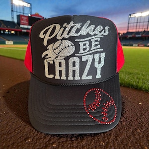 May include: Black baseball cap with red mesh panels. The cap displays the text "Pitches BE CRAZY" in silver glitter, accompanied by a baseball graphic. A baseball design made of red rhinestones is on the front.