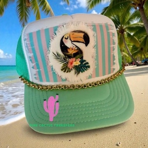 Könnte beinhalten: Grün-weiße Trucker-Cap mit Tukan- und Blumenmuster. Die Kappe hat eine rosa Kaktusgrafik auf dem Schirm und ein goldenes Kettendetail. Der Text "PINK CACTUS TRANSFERS" ist auf dem Schirm aufgedruckt.