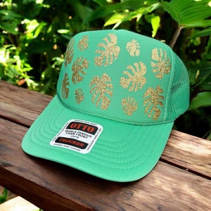 May include: A green trucker hat with a mesh back and a curved brim. The front of the hat features gold glitter monstera leaf designs. The hat has a white tag with black text that reads "OTTO WORLD FAMOUS FOAM FRONT TRUCKER".