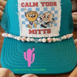 May include: Teal baseball cap with a mesh back and a beaded band. The front features a patch with cartoon baseball and glove characters and the text "CALM YOUR MITTS." A pink cactus graphic is below the patch. The text "PINK CACTUS TRANSFERS" is also visible.