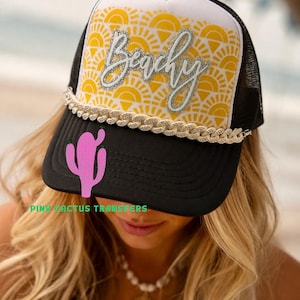 Op de afbeelding: Zwarte en witte truckerpet met een gele zonnepatroon en het woord "Beachy" in zilveren glitter. Een roze cactusafbeelding staat op de voorkant en een decoratieve touwrand siert de rand. Een zomeraccessoire.