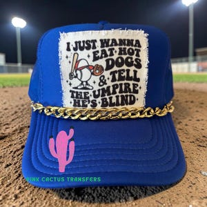 May include: A blue trucker hat with a gold chain and a pink cactus graphic. The hat features a patch with the text "I JUST WANNA EAT HOT DOGS & TELL THE UMPIRE HE'S BLIND" and a baseball-themed illustration.