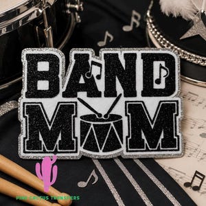 May include: A black glittery patch with the text BAND MOM, featuring a drum icon replacing the letter O. The letters have a silver glitter border. Musical notes are integrated into the design, which is set against a black and white background.