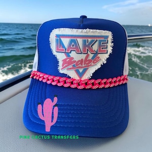 May include: A blue trucker hat with a white mesh back and a pink chain across the front. The hat features a patch with the words "LAKE Babe" in pink and blue, and a pink cactus graphic. The text "PINK CACTUS TRANSFERS" is at the bottom.