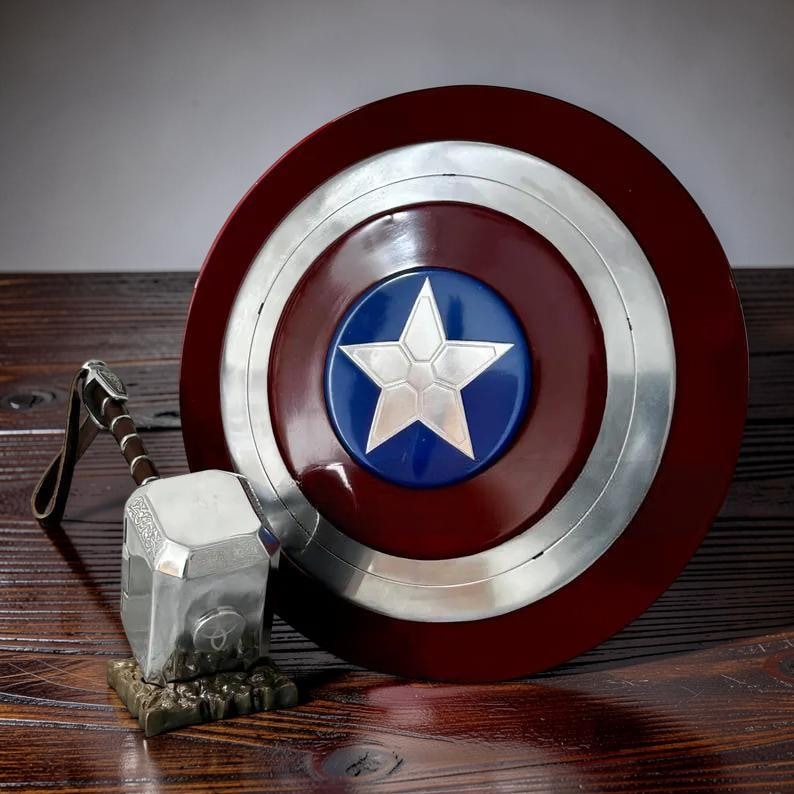 May include: A Captain America shield with a silver star on a blue circle, surrounded by a silver ring and a red outer ring. Next to it is a silver hammer with a brown handle, resting on a textured base. The items are on a wooden surface.