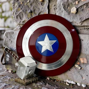 May include: A red, white, and blue Captain America shield with a silver star in the center. Next to the shield is a silver Thor's hammer with a brown leather strap. The shield and hammer are resting on a stone surface.