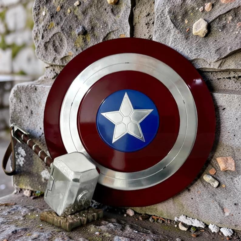 May include: A red, white, and blue Captain America shield with a silver star in the center. Next to the shield is a silver Thor's hammer with a brown leather strap. The shield and hammer are resting on a stone surface.