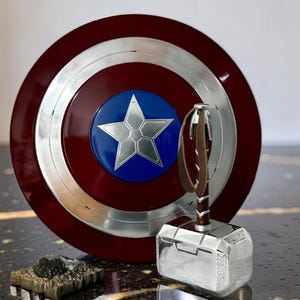 May include: A Captain America shield with a silver star on a blue circle, surrounded by a red and silver design. Next to it is Thor's hammer, Mjolnir, with a brown leather strap and a silver head. A small, detailed sculpture is in the foreground.