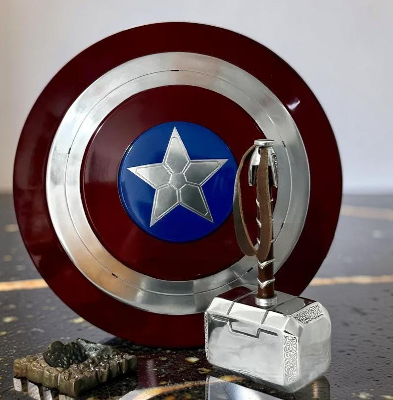 May include: A Captain America shield with a silver star on a blue circle, surrounded by a red and silver design. Next to it is Thor's hammer, Mjolnir, with a brown leather strap and a silver head. A small, detailed sculpture is in the foreground.