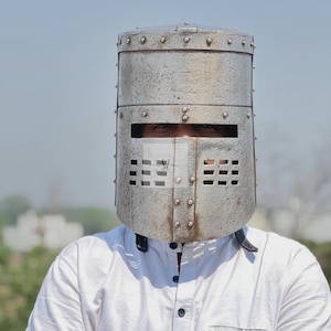 May include: A riveted metal knight's helmet with rectangular eye and mouth openings. The helmet is cylindrical and appears to be made of heavy steel. The person wearing the helmet is wearing a white button-down shirt.