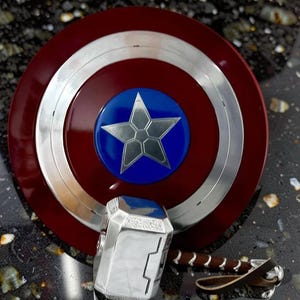 May include: A Captain America shield with a silver star on a blue circle in the center, surrounded by a silver ring and a red outer ring. A silver hammer with a brown leather strap is also present.