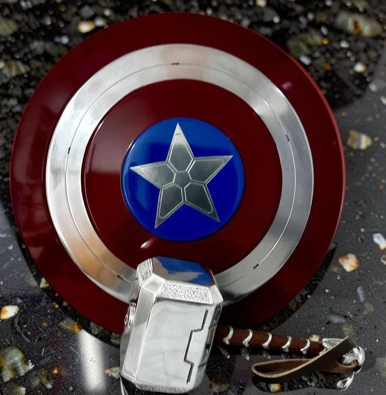 May include: A Captain America shield with a silver star on a blue circle in the center, surrounded by a silver ring and a red outer ring. A silver hammer with a brown leather strap is also present.
