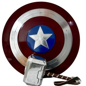 May include: A Captain America shield with a silver star on a blue circle, surrounded by a silver ring and a red background. A silver hammer with a brown leather strap rests below the shield.