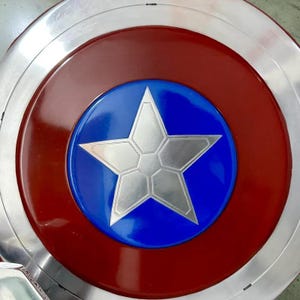 May include: A metallic shield with a red and silver outer ring, a red inner ring, and a blue center. A large, silver, five-pointed star is in the center of the blue circle. The shield is a replica of a superhero's shield.