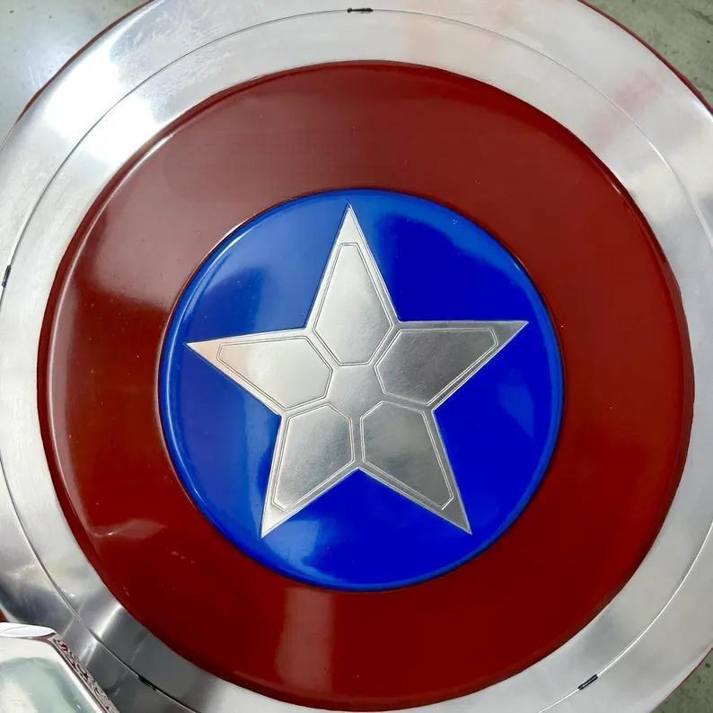 May include: A metallic shield with a red and silver outer ring, a red inner ring, and a blue center. A large, silver, five-pointed star is in the center of the blue circle. The shield is a replica of a superhero's shield.