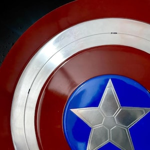 May include: A close-up of a Captain America shield. The shield features a red and white striped outer ring, a silver metallic inner ring, and a blue center with a silver star. The shield has a glossy finish.