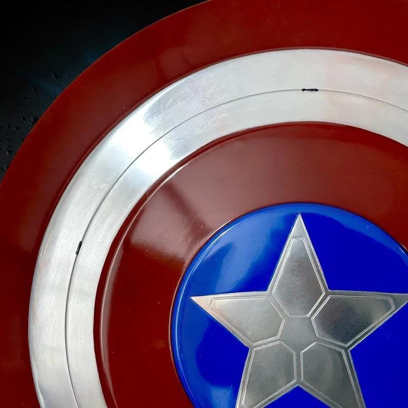 May include: A close-up of a Captain America shield. The shield features a red and white striped outer ring, a silver metallic inner ring, and a blue center with a silver star. The shield has a glossy finish.