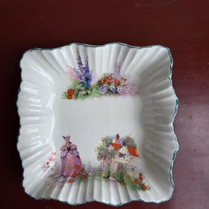 May include: A white, square dish with a ruffled edge and a painted landscape scene. The scene shows a woman in a pink dress, a cottage with an orange roof, and flowers. The rim has a light blue trim.