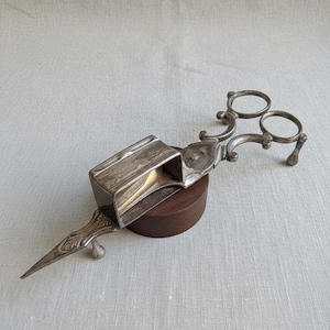 May include: An antique silver-coloured scissor-like object with a rectangular box and two finger rings. The object has a pointed end and rests on a small, round, wooden base. The design features ornate detailing.