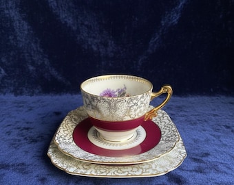 May include: A vintage teacup, saucer, and plate set. The teacup has a gold handle and rim, with a floral design inside. The saucer and plate are white with a gold filigree pattern and a burgundy center. The set is on a blue background.