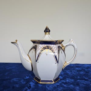 May include: White porcelain teapot with a geometric design, accented with gold trim and navy blue details. The lid is topped with a gold and navy finial. The teapot has a curved spout and a handle. A classic piece for tea time.