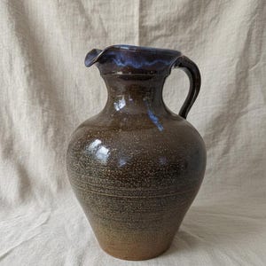 May include: A handmade ceramic jug with a dark brown exterior and a blue interior rim. The jug has a handle and a ruffled edge. The pottery has a textured surface and a rustic appearance, suitable for serving drinks.