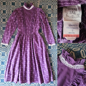 May include: A vintage purple Gunne Sax dress with a floral pattern. The dress has long sleeves, a high collar, and white lace trim. The label reads "Gunne Sax by Jessica McClintock."