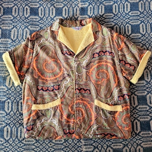 May include: A short-sleeved button-down shirt with a paisley pattern in shades of brown, orange, and green. The shirt has a yellow collar, cuffs, and pocket trim. The shirt is lying on a blue patterned fabric.
