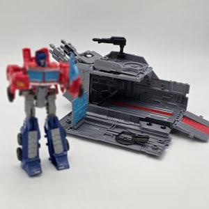 May include: A red, blue, and grey robot action figure stands beside a grey transport vehicle with a black gun turret. The vehicle has a red interior and a blue door. The robot has blue legs and a blue chest.