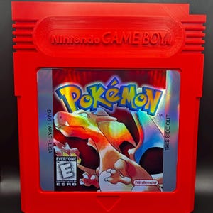 May include: A red Game Boy cartridge featuring the Pokémon game. The cartridge has the Nintendo Game Boy logo embossed on the top. The game's label displays the Pokémon logo and a Charizard illustration. The ESRB rating is visible.