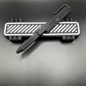 May include: A black pen with the text "P365" on the side rests on a black and white rectangular base. The pen has a futuristic design with a clip and a textured grip. The base has a white striped pattern.