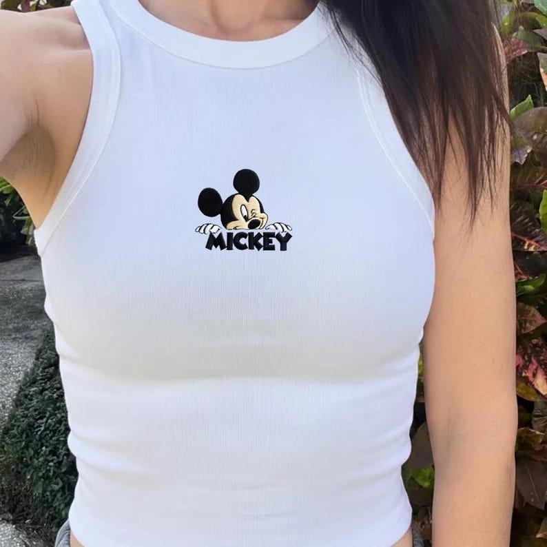May include: White ribbed vest top with a Mickey Mouse graphic. The graphic shows Mickey Mouse's face and the word "MICKEY" in black. The vest top has a scoop neck and is sleeveless.