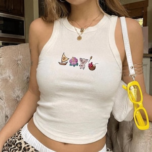 Op de afbeelding: Cr&egrave;mekleurige tanktop met geborduurde cartoonafbeeldingen van een boot, bloem, varken en kokosnoot. De top heeft een ronde halslijn en een mouwloos ontwerp. Het model draagt gouden kettingen en een witte handtas met gele zonnebril.