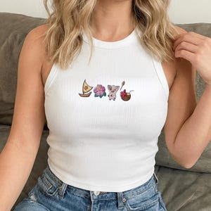 Op de afbeelding: Witte geribbelde tanktop met geborduurde cartoonafbeeldingen van een zeilboot, bloem, varken en kokosnoot. De mouwloze top heeft een ronde halslijn. Het ontwerp is in tinten bruin, roze en paars.