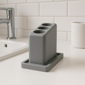 May include: A gray toothbrush holder with three circular openings. The holder is rectangular with a small tray at the base. The holder is made of a solid, matte material and sits on a white countertop. A white ceramic cup is in the background.