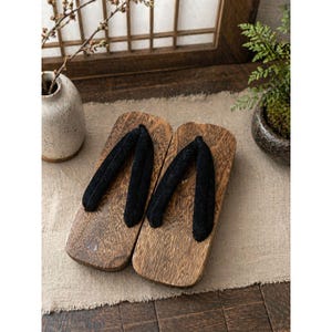 May include: A pair of traditional Japanese geta sandals. The wooden sandals have a natural wood grain finish and are paired with black fabric straps. The sandals are displayed on a neutral-colored cloth, with a blurred background featuring a vase and a small potted plant.