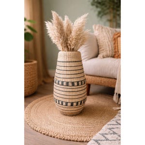 May include: A tall, woven vase with a black and natural color pattern, filled with fluffy, beige pampas grass. The vase sits on a round, woven jute rug. The background includes a sofa and other decorative elements.