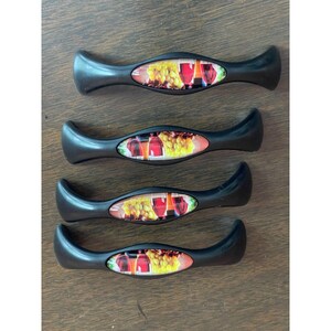 May include: Four black cabinet pulls with a curved, elongated shape. Each handle features a central oval inlay displaying a colorful graphic of red wine glasses and clusters of green grapes against a light background, set within the matte black frame.