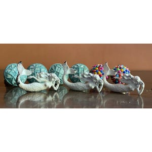 May include: Three white mermaid figurines are arranged in a row, each featuring a curved tail and flowing hair. Behind the mermaids are several decorative spheres, including two textured teal globes and two colorful, multi-beaded round ornaments.