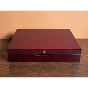 May include: A rectangular, dark burgundy wooden box with a smooth, polished finish. The box features a hinged lid and a small, centered silver-toned metal knob on the front edge for opening. The design is simple, flat, and solid in construction.