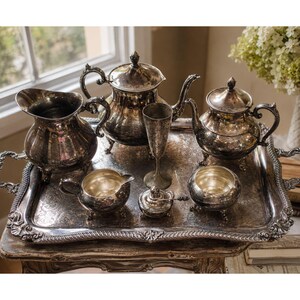 May include: A silver-plated tea service set on a rectangular tray, including a large teapot, a smaller teapot, a creamer, a sugar bowl with a lid, a pitcher, and a tall stemmed goblet, all featuring ornate embossed patterns and dark patina.
