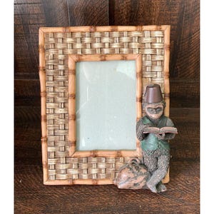 May include: A rectangular picture frame featuring a woven light brown texture with bamboo-style borders. A sculpted figure of a monkey wearing a green vest and a brown fez hat stands on the bottom right, holding an open book with a brown bag nearby.