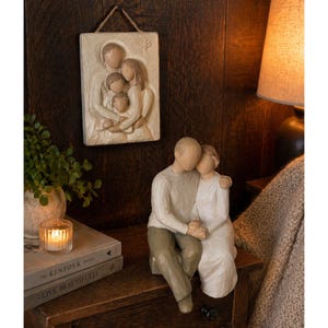 Könnte beinhalten: Ein familienorientiertes Dekorationsset mit einer Relief-Wandplakette und einer sitzenden Paarfigur. Die Wandplakette zeigt eine vierköpfige Familie, die sich umarmt. Die Figur zeigt ein sich umarmendes Paar. Beide sind in neutralen Tönen gehalten.