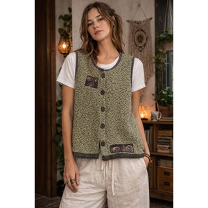 May include: A green knitted vest with a button-up front, featuring decorative patches and a dark gray trim. The vest is worn over a white t-shirt and paired with light-colored pants. The style is casual and bohemian.