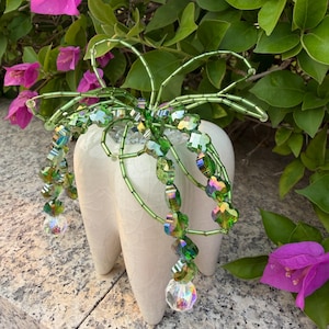 Pode incluir: Um vaso branco de cerâmica em forma de dente com um design floral de contas verdes. O design de contas apresenta contas verdes e cristais iridescentes. O fundo inclui folhas verdes e flores de buganvílias cor-de-rosa.